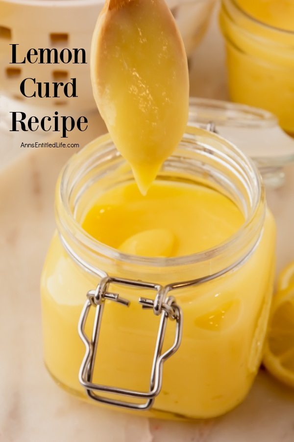 Lemon curd clinging to a spoon held above a jar of homemade lemon curd. There is a second jar in the upper right, a basket of lemons in the upper left.