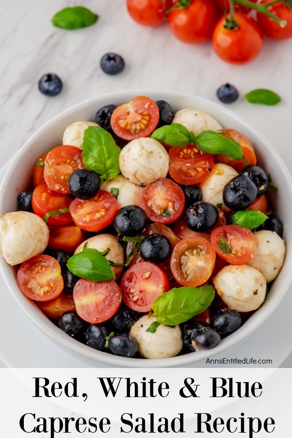An overhead view of the mozzarella, blueberries, basil, and balsamic glaze in a red, white, and blue Caprese salad in a white bowl. There are blueberries, fresh basil, and cherry tomatoes above the bowl.
