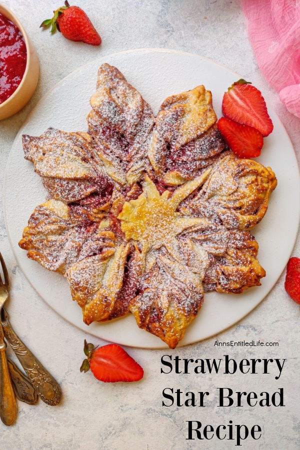 Overhead view of a strawberry bread in the shape of a star on a white plate. Fresh, sliced strawberries garnish the plate, gold silverware is in the lower left, a cup of strawberry jam is in the upper left.