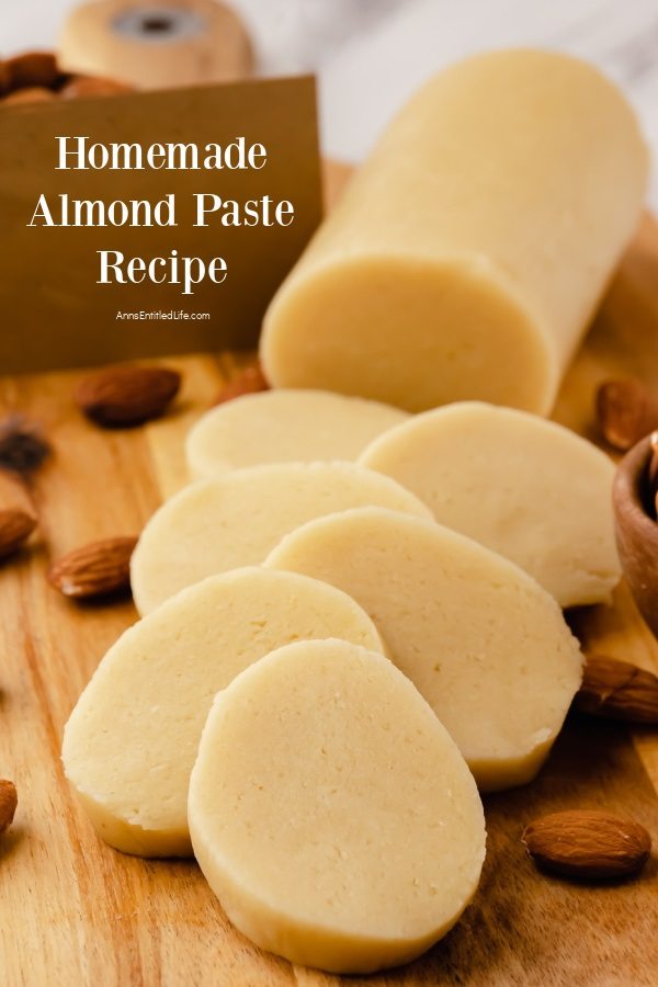 A roll of homemade almond paste on a wood cutting board surrounded by almonds, slices are stacked in from of the roll.