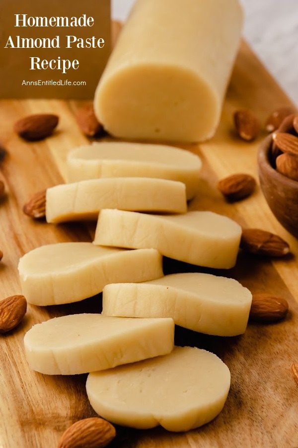 Head-on view of a roll of homemade almond paste on a wood cutting board surrounded by almonds, slices are stacked in from of the roll.