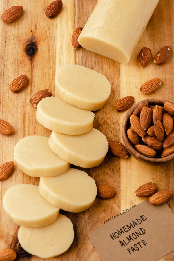 Overhead view of roll of homemade almond paste on a wood cutting board surrounded by almonds, slices are stacked in from of the roll, and a small wooden bowl to the right is filled with almonds.