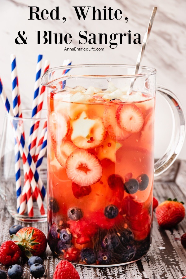 A side view of a pitcher of red, white, and blue sangria is a clear pitcher. There are red and white and blue and white straws in a glass to the right.