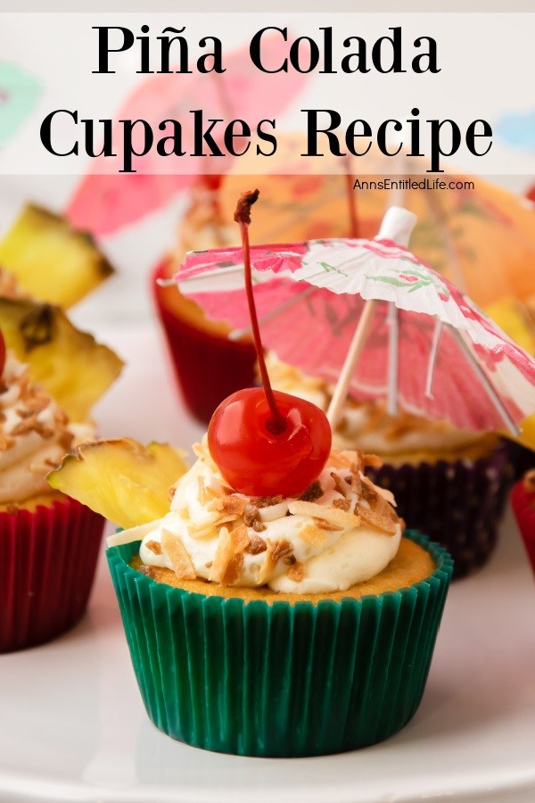A pina colada cupcake in a green cupcake liner frosted and garnished with pineapple, toasted coconut, and a paper umbrella is front and center in this photo. There are other pina colada cupcakes in the background.