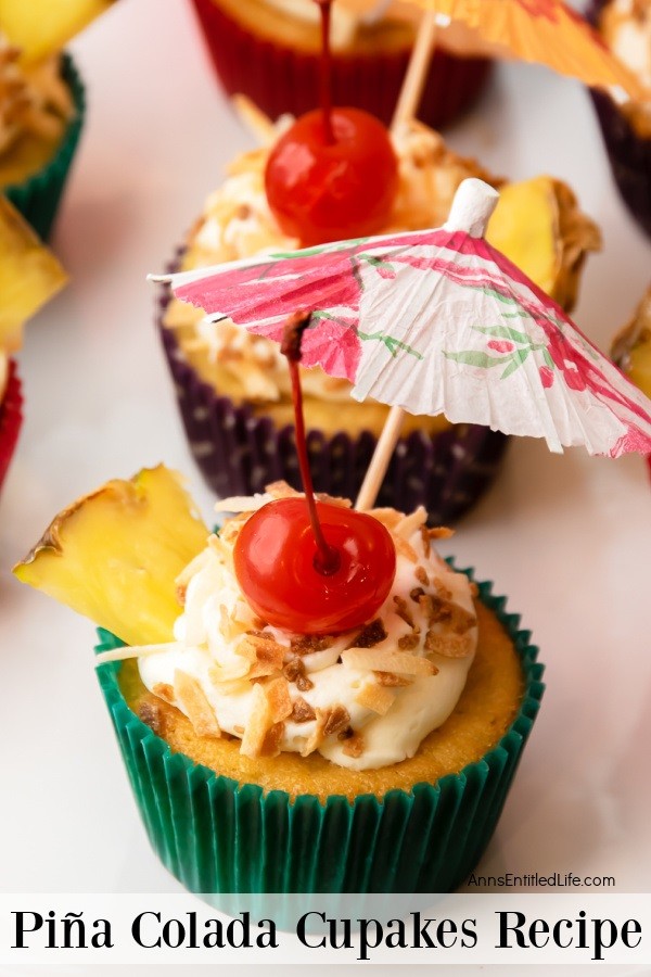 An overhead view of a  pina colada cupcake in a green cupcake liner frosted and garnished with pineapple, toasted coconut, and a paper umbrella sitting front and center in this photo. There are other pina colada cupcakes in the background.