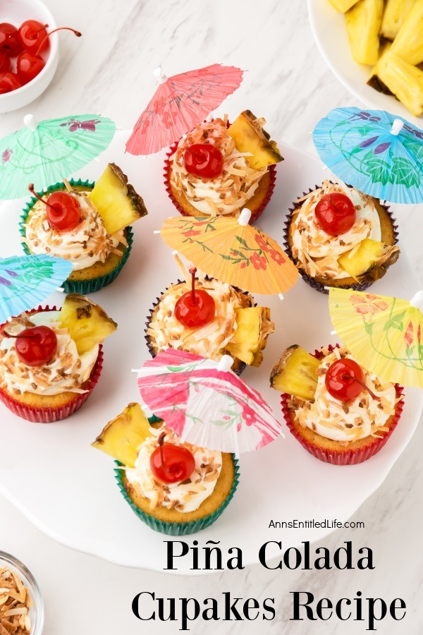 An overhead view of a white plate filled with pina colada cupcakes garnished with pineapple, toasted coconut, and paper umbrella. There is a white bowl filled with cut pineapple in the upper right, a bowl of maraschino cherries in the upper left.