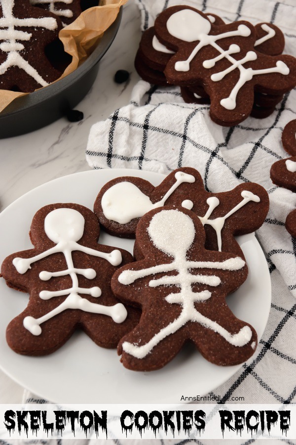 A white plate filled with three chocolate cookies in a skeleton shape with skeleton decorations on a black and white checked dishcloth. A bowl filled with more cookies in the upper left, a stack of cookies is in the upper right.