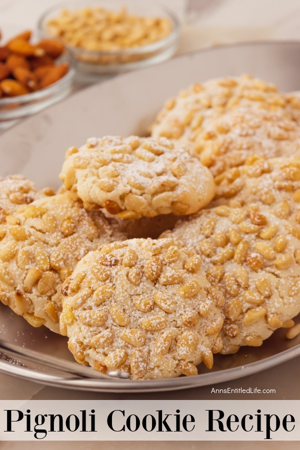 Close-up photo of an of a white platter filled with pignoli cookies, a bowl of nuts and a bowl of pine nuts is in the upper left