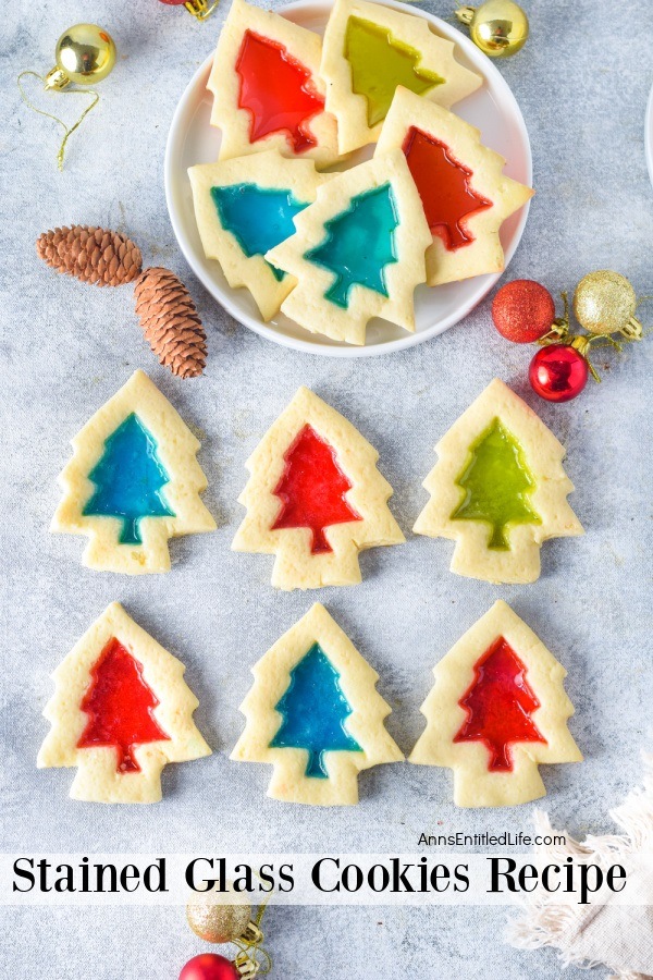 Six stained glass cookies laid out on a blue countertop, a white plate of cookies is in the upper center. Small holiday decorations surround the cookies.
