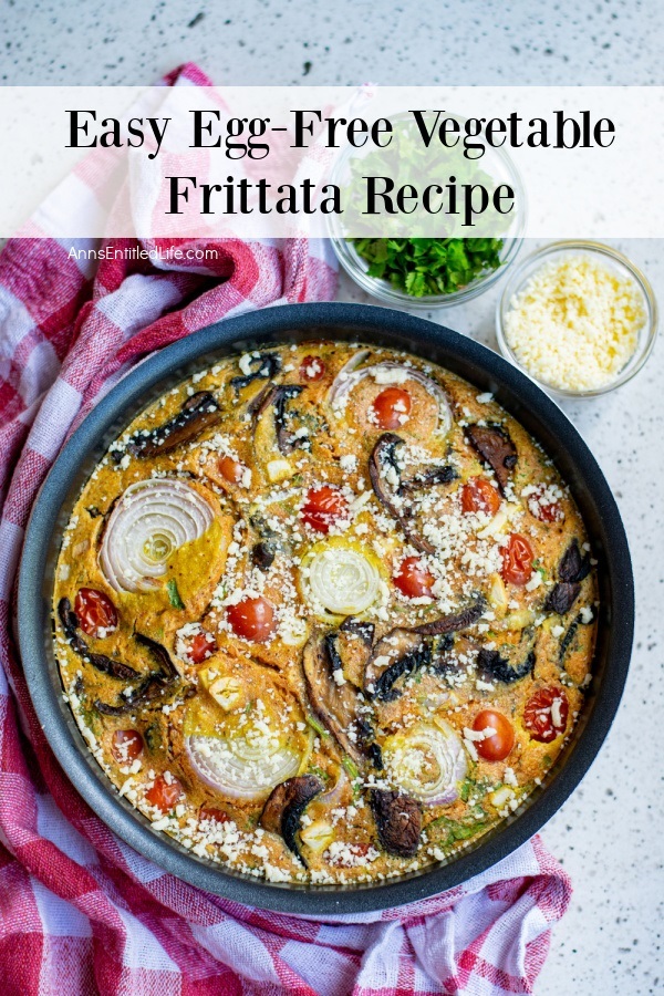 a skillet baked with egg-free vegetable frittata sitting on a grey counter, there is a small container of cheese on the right, and shredded cilantro on the upper right, a red and white checked napkin on the left