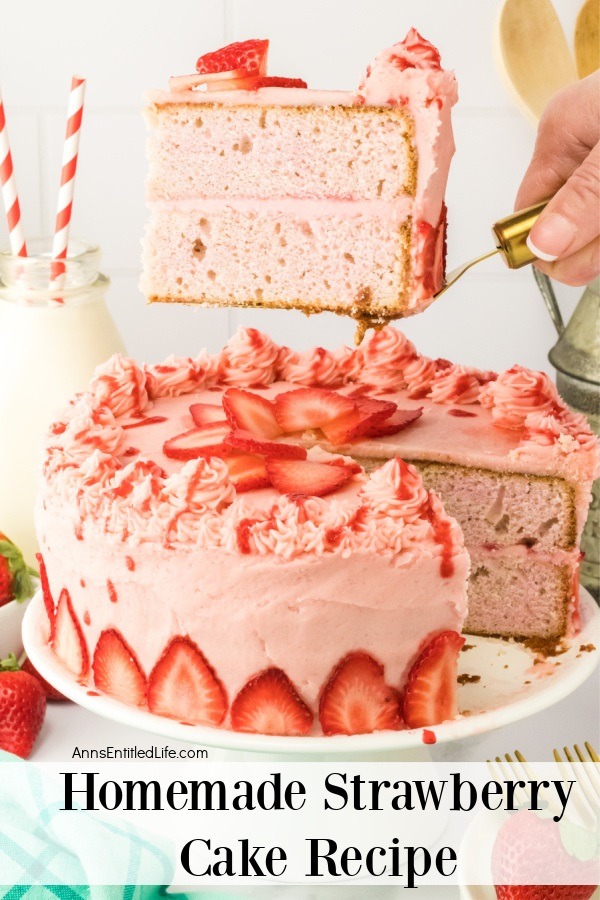 A piece of strawberry cake is being lifted on a gold cake server above a strawberry cake on a white cake stand. the is a milk jug in the upper left.