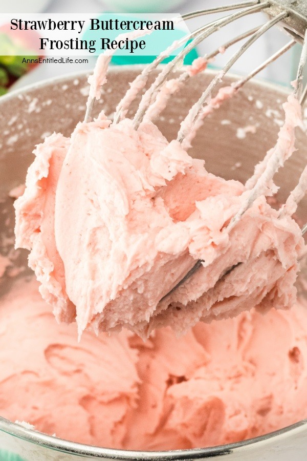 A close-up photo of a mixing bowl filled with strawberry frosting, the whisk is raised and covered with strawberry frosting