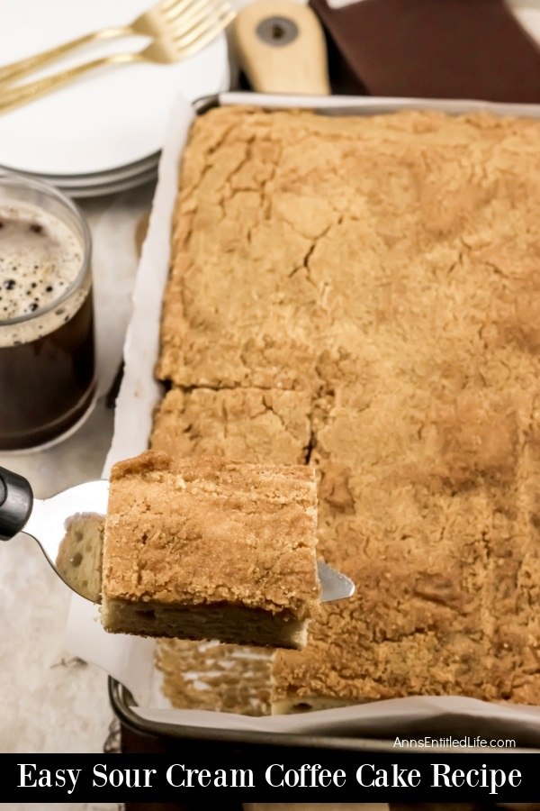 A baking pan filled with sour cream coffee cake, one piece is being lifted out of the pan. There are a stack of white plates and a cup of black coffee to the left of the pan
