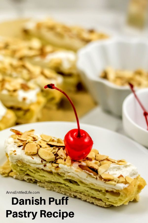 side view of a piece of Danish pastry puff on a white plate surrounded by the rest of the Danish puff and a bowl of almonds