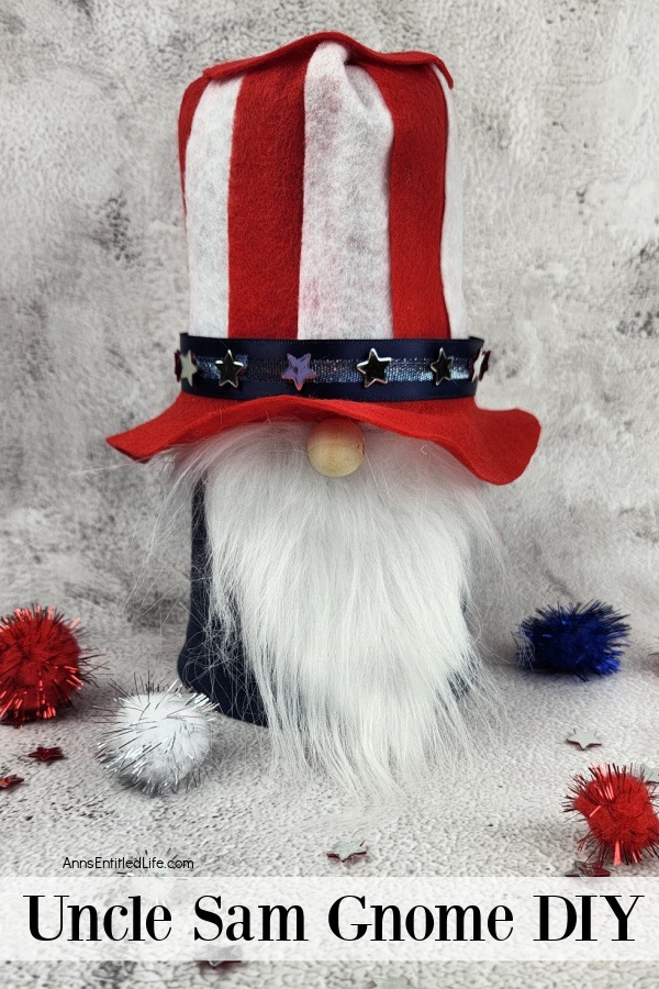 a homemade patriotic gnome that resembles Uncle Sam against a marble top and background, a few red, white, and blue pom poms are scattered at its bottom