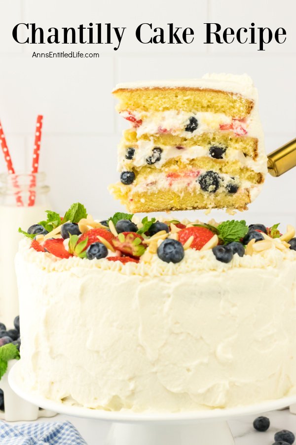 A chantilly cake on a white cake dish, one piece is being lifted by a cake lifter. There is a glass of milk in the upper left.