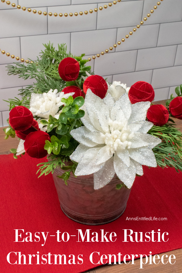 A rustic farmhouse style faux floral holiday arrangement in a tin bucket sits atop a red place mat against a white brick counter with a gold bead garland strung behind the flowers.