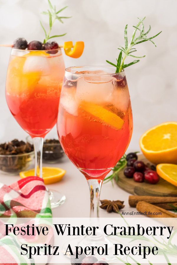 Two wine glasses full of cranberry spritz Aperol decorated with cranberries, rosemary sprig, and orange curl surrounded by a cutting board of cut oranges and cranberries as well as cinnamon sticks in the upper right. There are clear bowls full of spices in the upper left.