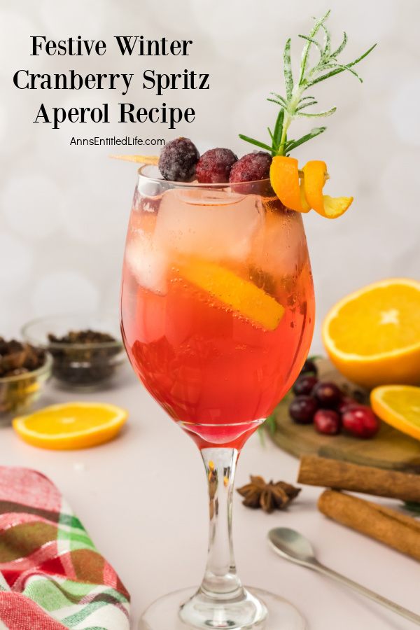 A wine glass of cranberry spritz Aperol decorated with cranberries, rosemary sprig, and orange curl surrounded by a cutting board of cut oranges and cranberries as well as cinnamon sticks in the upper right. There are clear bowls full of spices in the upper left.