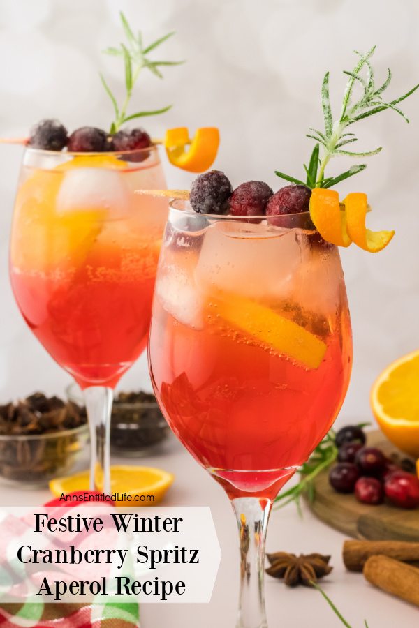 A close up of two wine glasses full of cranberry spritz Aperol decorated with cranberries, rosemary sprig, and orange curl surrounded by a cutting board of cut oranges and cranberries as well as cinnamon sticks in the upper right. There are clear bowls full of spices in the upper left.