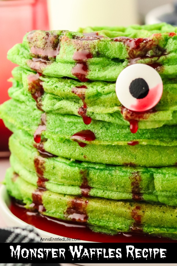 A close up of a stack of green waffles drenched in red syrup on a white plate. The waffles are topped with berries and candy eyes.