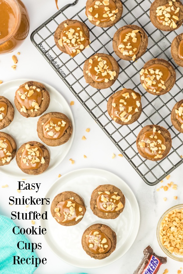 10 snickers stuffed cookie cups on a wire rack in the upper right, five on a white plate on the left, three on a white plate center bottom. There is a Snickers bar in the lower right, a clear bowl filled with chopped peanuts in the lower right.