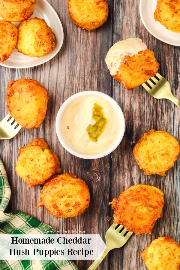 Cheddar hush puppies on a wooden platter surrounding a white bowl filled with chili aioli, several hush puppies dipped in the sauce are on a fork 