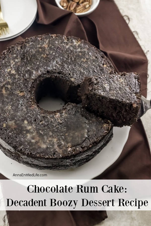 An overhead view of a chocolate rum cake on a white plate, there is a piece being lifted on a cake lifter. There is a stack of white plates above it, along with a white bowl of pecans.