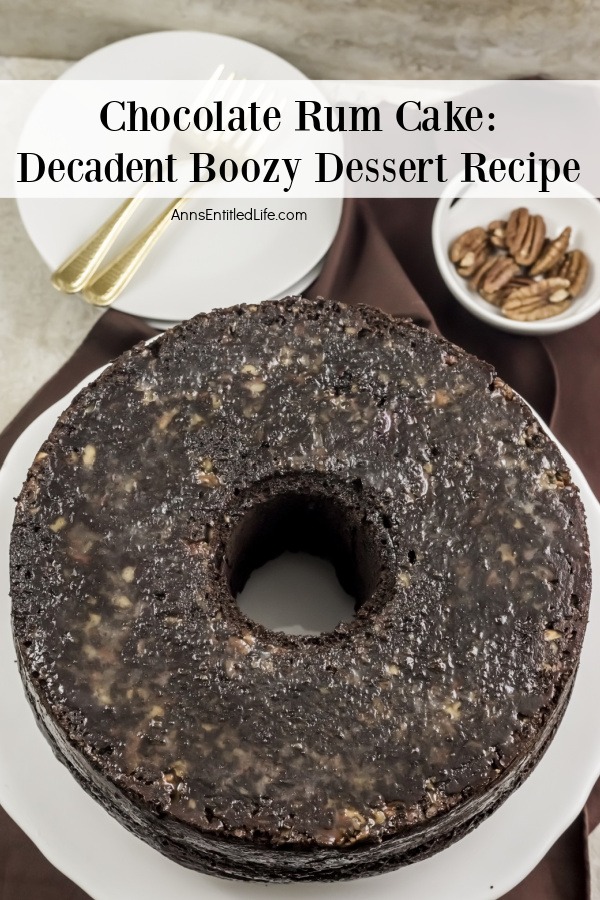 An overhead view of a whole chocolate rum cake on a white plate. There is a stack of white plates above it, along with a white bowl of pecans.