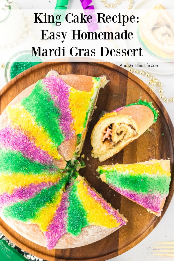 An overhead view of a whole king cake on a wooden server, a piece is turned on its side to see the baby, another cut piece is pulled away from the rest of the cake. There are beads and colored sugar above the platter.
