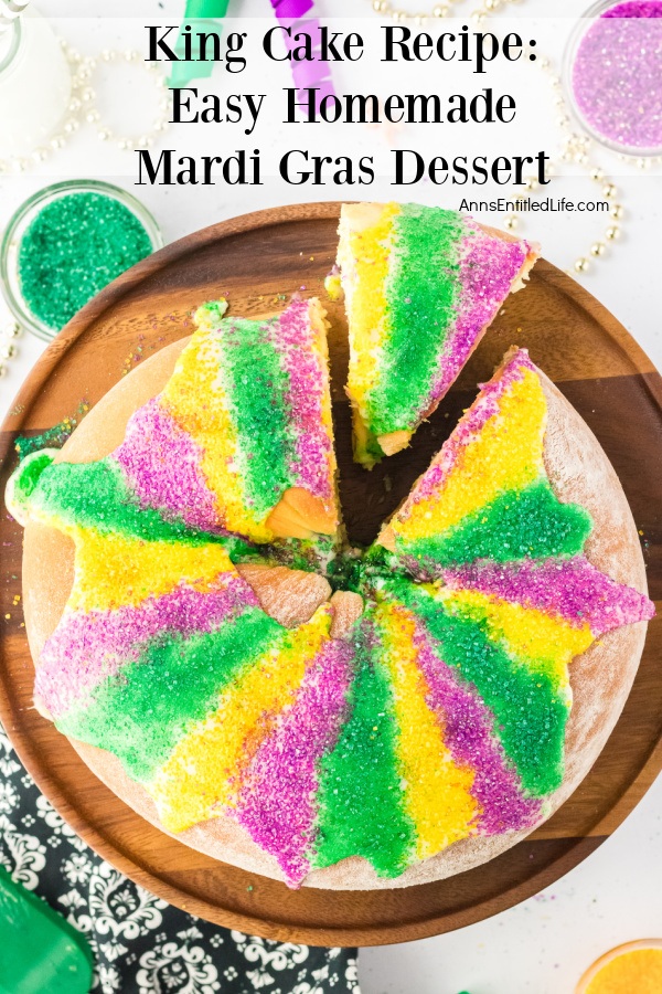 An overhead view of a whole king cake on a wooden server, there is one piece cut away from the rest of the king cake. There are beads and colored sugar above the platter.