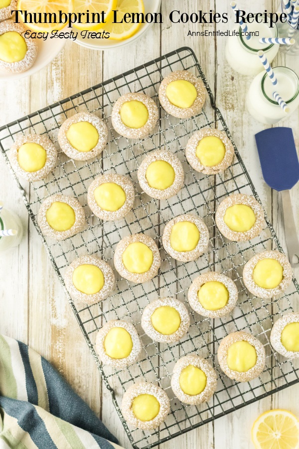 Thumbprint lemon cookies on a wire rack atop a rustic board. Milk bottles are in the upper right, a rubber spatula below them. Bowls of cut lemon slices and a white plate filled with thumbprint lemon cookies is in the upper left.