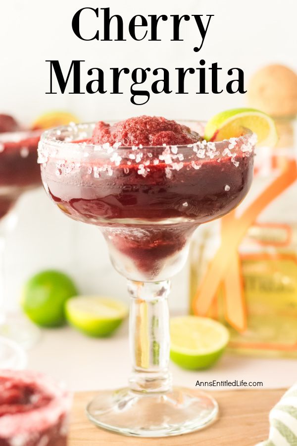 A frozen cherry margarita in a margarita glass on a cutting board. Background contains a small cocktail glass filled with cherry margarita, some cut limes, and a bottle of tequila